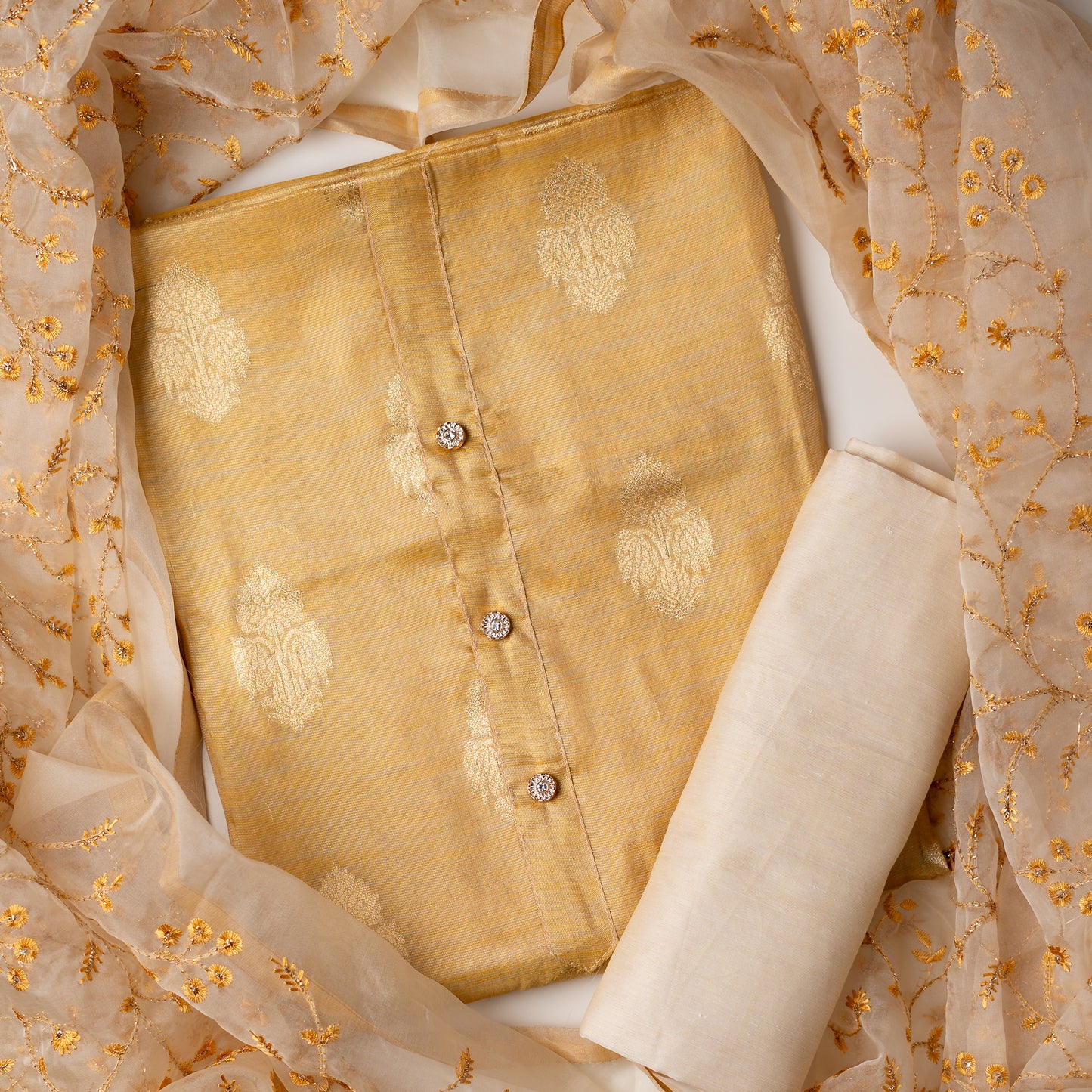 An elegantly crafted Tissue Silk Party wear dress material. A set best suited to gift bride to be or your loved on your anniversary. Tissue silk top with golden color embossed designs, it has show buttons in neck area, also golden color borders in side. Silk dupatta with embroidery work with the same color thread of top and also golden color threads. Cream color cotton silk bottom.