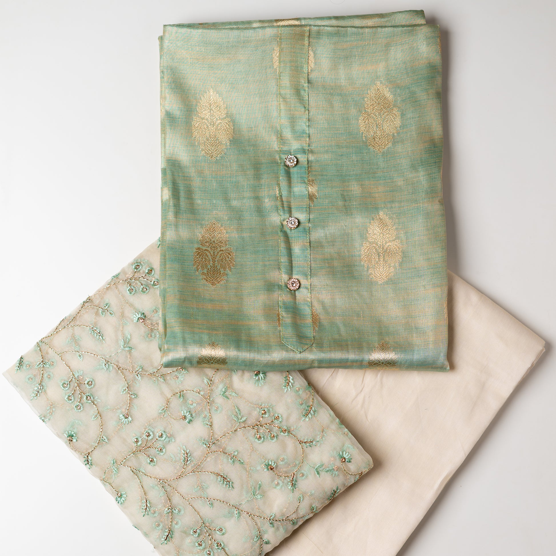 An elegantly crafted Tissue Silk Party wear dress material. A set best suited to gift bride to be or your loved on your anniversary. Tissue silk top with golden color embossed designs, it has show buttons in neck area, also golden color borders in side. Tissue silk dupatta with embroidery work with the same color thread of top and also golden color threads. Cream color cotton silk bottom.