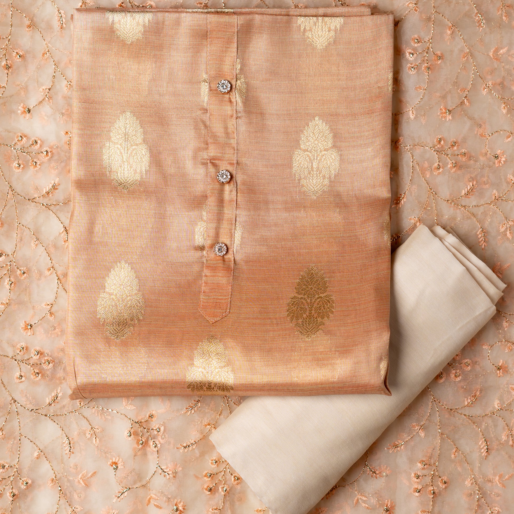 An elegantly crafted Tissue Silk Party wear dress material. A set best suited to gift bride to be or your loved on your anniversary. Tissue silk top with golden color embossed designs, it has show buttons in neck area, also golden color borders in side. Tissue silk dupatta with embroidery work with the same color thread of top and also golden color threads. Cream color cotton silk bottom.