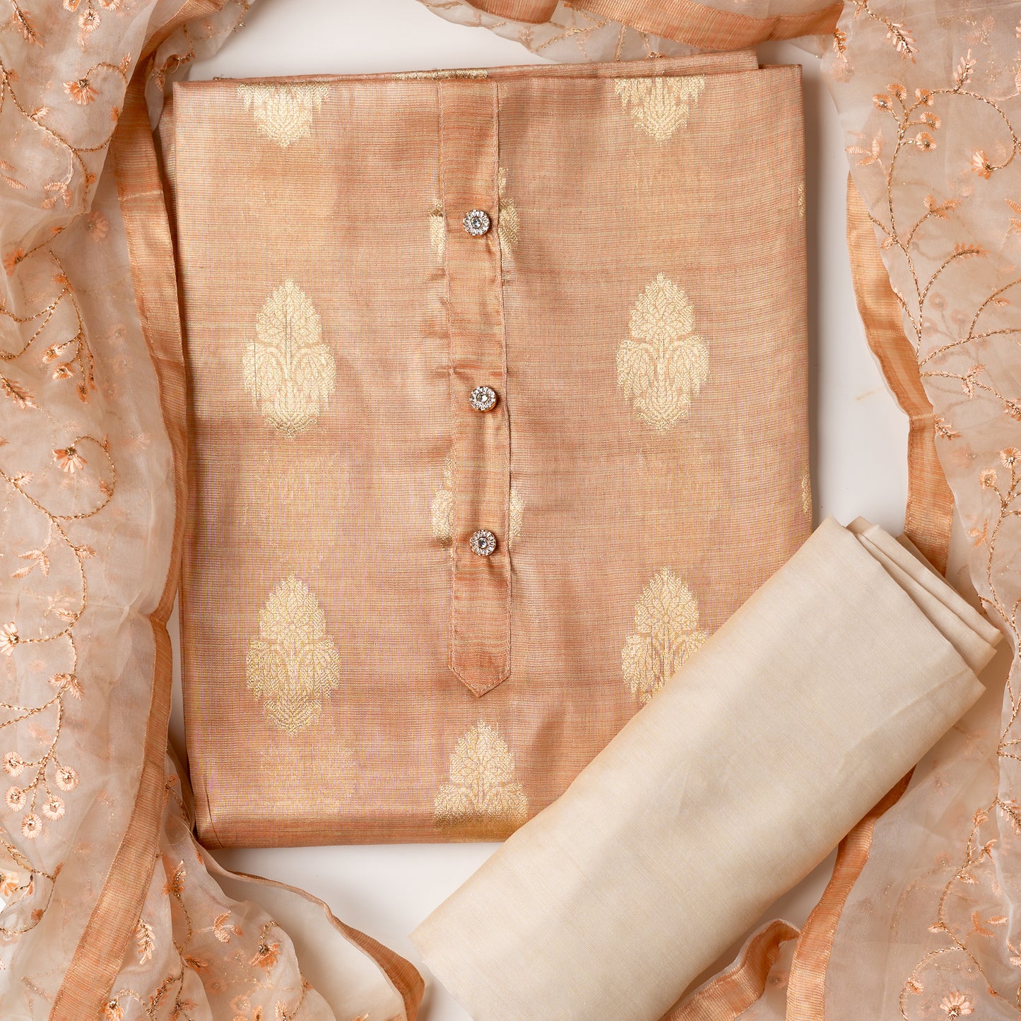 An elegantly crafted Tissue Silk Party wear dress material. A set best suited to gift bride to be or your loved on your anniversary. Tissue silk top with golden color embossed designs, it has show buttons in neck area, also golden color borders in side. Tissue silk dupatta with embroidery work with the same color thread of top and also golden color threads. Cream color cotton silk bottom. 