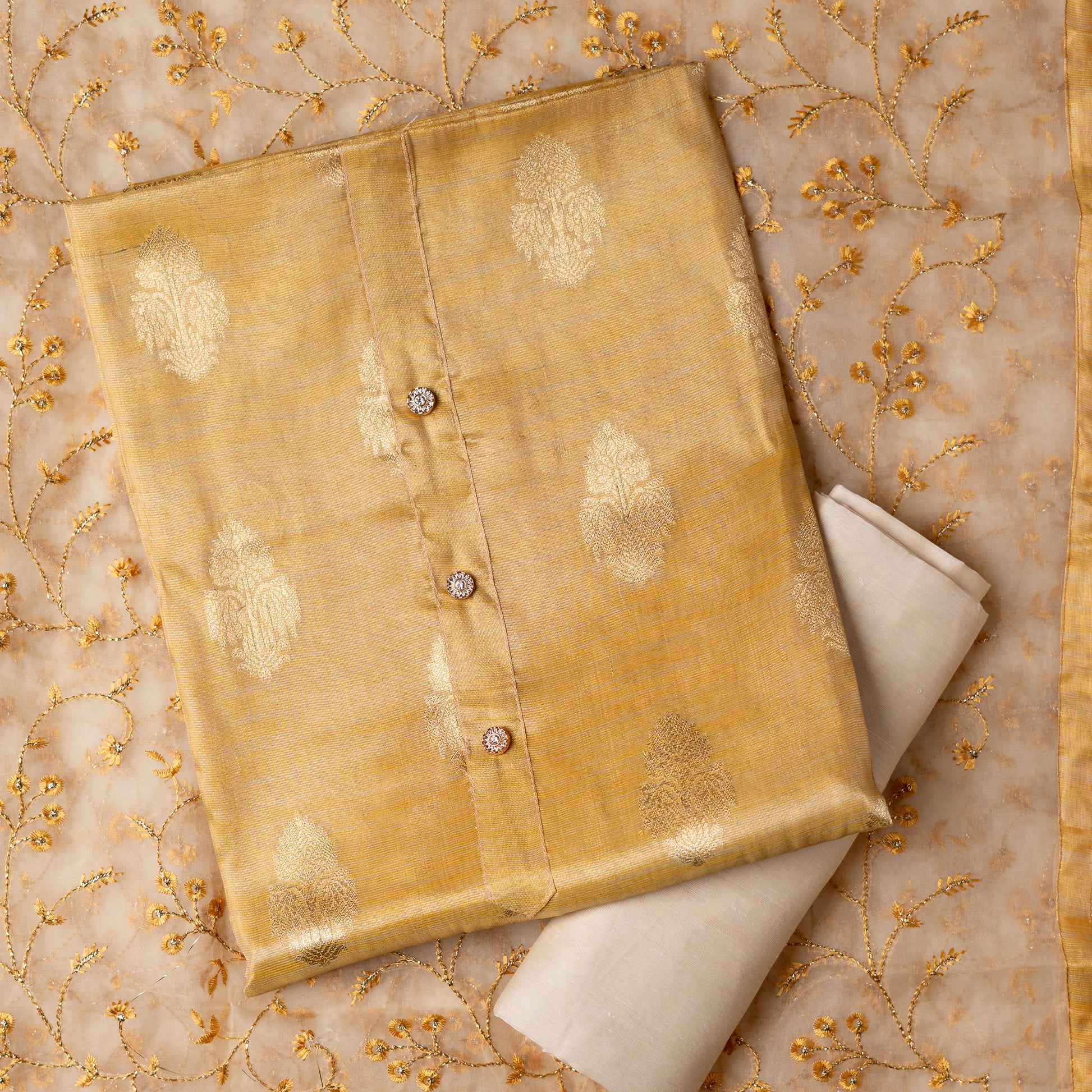 An elegantly crafted Tissue Silk Party wear dress material. A set best suited to gift bride to be or your loved on your anniversary. Tissue silk top with golden color embossed designs, it has show buttons in neck area, also golden color borders in side. Silk dupatta with embroidery work with the same color thread of top and also golden color threads. Cream color cotton silk bottom. 