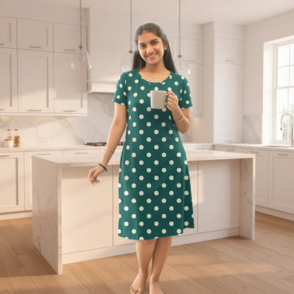 A woman stands in a modern kitchen wearing a teal green short-sleeve nightgown with white polka dots and holding a white mug.