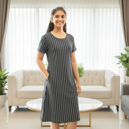 A woman stands in a bright living room wearing a short-sleeve knee-length dress with thin vertical white pinstripes on a dark gray background. The dress has side pockets and a round neckline.