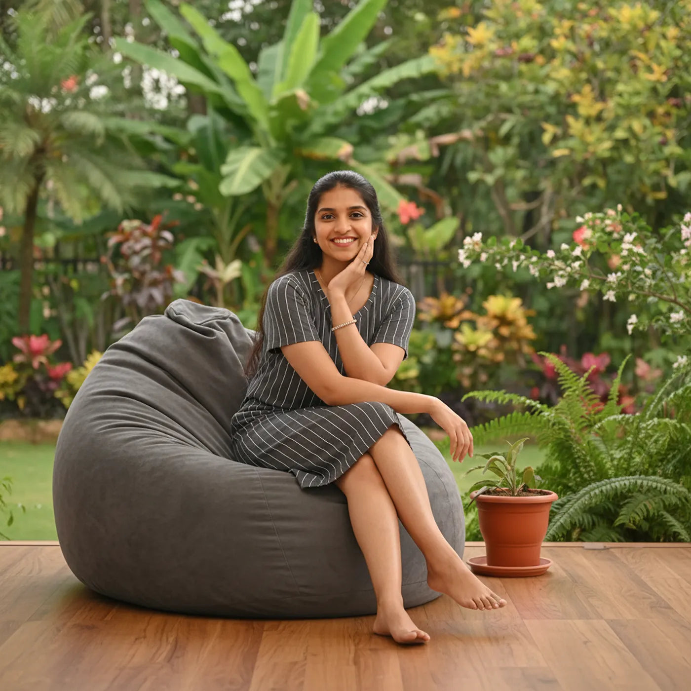 A woman wearing a grey stripe design lounge dress and sits barefoot on a large dark gray bean bag chair on a wooden deck, with a lush tropical garden background.