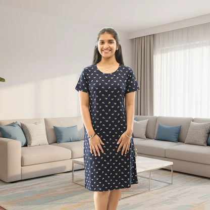 A woman stands in a modern living room wearing a navy blue cotton knee length short nighty  with a small white floral pattern. 