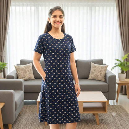 A woman stands in a bright living room wearing a navy blue cotton knee length short nighty with pocket, it has print design all over with a small white floral pattern. 