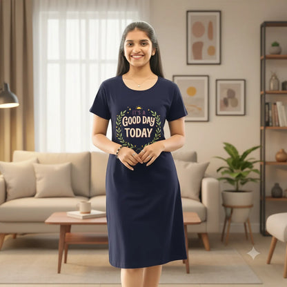 A woman stands in a cozy living room wearing a navy blue hosiery cotton knee length short nighty with a 'Good Day TODAY' graphic on the front. The room features a light gray sofa with patterned pillows, potted plants, and warm lighting from floor lamps.