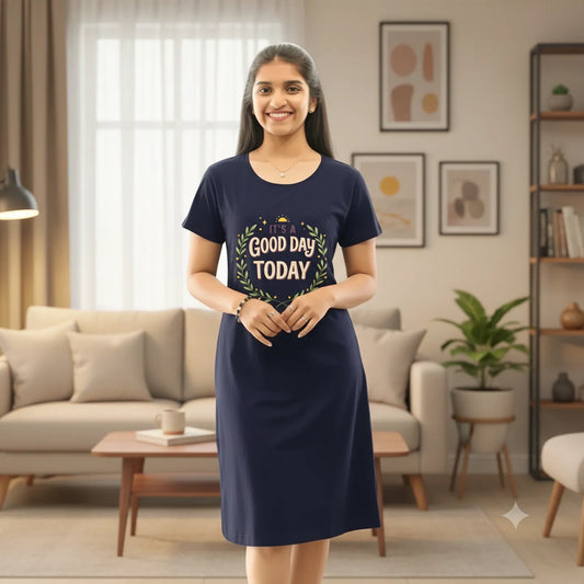 A woman stands in a cozy living room wearing a navy blue hosiery cotton knee length short nighty with a 'Good Day TODAY' graphic on the front. The room features a light gray sofa with patterned pillows, potted plants, and warm lighting from floor lamps.