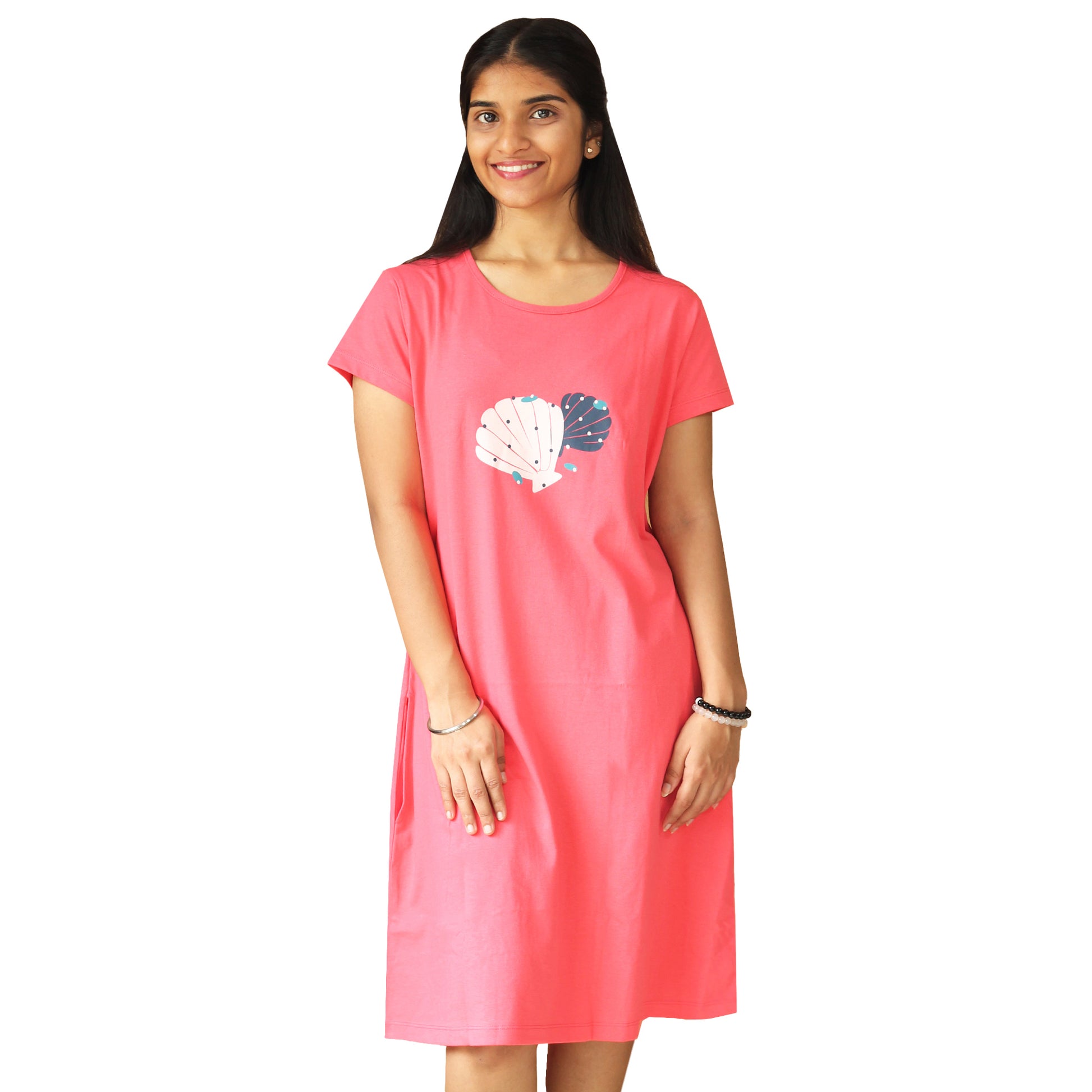 A woman standing and wearing a knee-length coral pink T-shirt dress with short sleeves and a round neckline. The dress features a shell print.