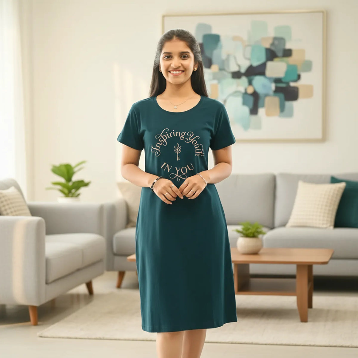 A woman stands in a living room wearing a dark green cotton short nighty  with a gold circular graphic on the front. 