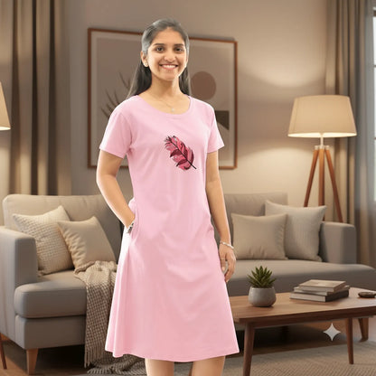 A woman wearing a light pink cotton knee length short  nighty with a red and pink feather graphic on the front, standing in a bright, modern living room with a gray sofa and plants.