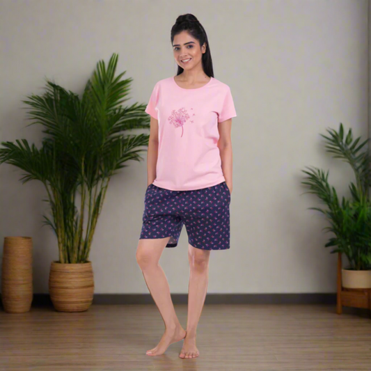 Woman wearing a pink t-shirt and dark blue patterned shorts standing in a room with plants.
